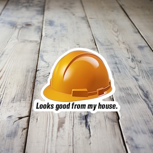 May include: A yellow hard hat with a black strap and the text "Looks good from my house." on a wooden surface.