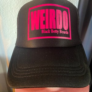May include: Black trucker hat with a pink square that says "WEIRDO" in bold letters. Below the word "WEIRDO" is the text "Black Betty Boards". The hat has a curved brim and a mesh back.