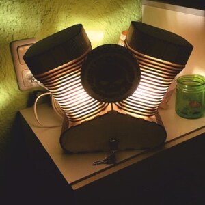 Pode incluir: Uma lâmpada única com design de motor de motocicleta. A lâmpada apresenta dois abajures cilíndricos com design canelado, um elemento central circular e uma base. Os abajures são pretos no topo e emitem luz quente. Uma chave está colocada na base.