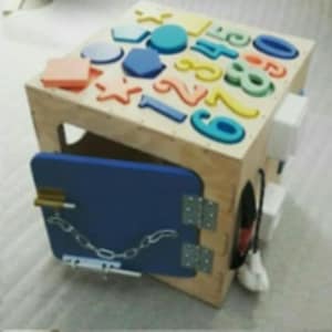 Può includere: Cubo attività in legno con forme, numeri e una porta blu con catena. Il cubo ha vari elementi interattivi per l'apprendimento e lo sviluppo precoce. La parte superiore del cubo presenta forme e numeri colorati.