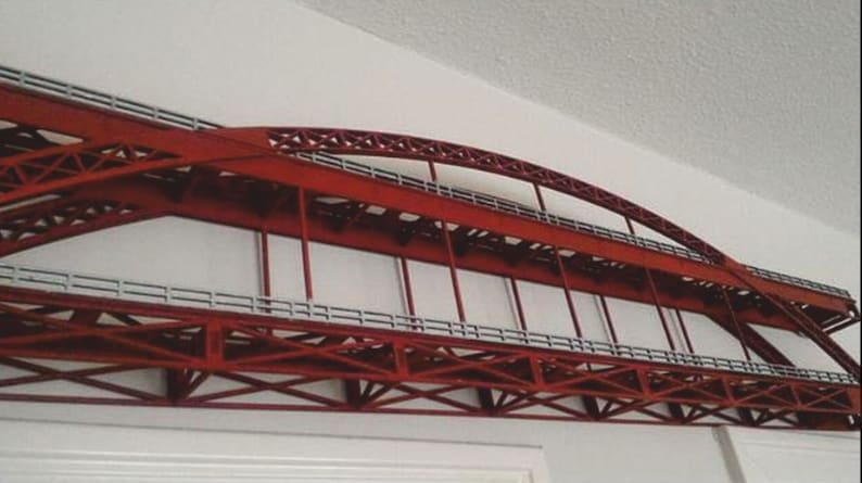 May include: A red metal model bridge with a detailed arch design. The bridge has a gray railing and is suspended from the ceiling.