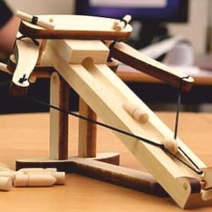 May include: A wooden catapult toy with a rubber band and a wooden arm. The catapult is made of light wood and has a dark brown base. The catapult is loaded with small wooden projectiles.