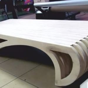 Könnte beinhalten: Moderner Couchtisch aus hellem Holz. Der Tisch hat eine rechteckige Oberseite mit Lamellendesign und geschwungenen, offenen Seiten. Der Tisch ist ca. 122 cm lang und 61 cm breit.