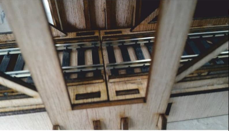 May include: A wooden model train bridge with two sets of tracks. The bridge is made of light brown wood and has a natural wood finish. The tracks are made of black plastic and are laid out in a straight line.