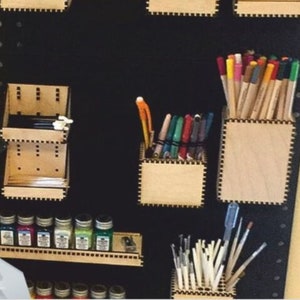 May include: A black pegboard with various wooden storage containers for art supplies. The containers hold colored pencils, markers, paint bottles, and paintbrushes.
