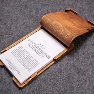 Może przedstawiać: Drewniany notes z unikalnym wzorem. Okładka wykonana jest z drewna i ma naturalne wykończenie. Notes jest otwarty, ukazując pustą stronę z nadrukowanym tekstem "Это деревянный блокнот".