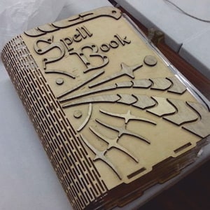May include: A wooden spell book with the words "Spell Book" carved into the cover. The cover features star and feather designs. The book's spine has a detailed, slotted design. The book is made of light-colored wood.