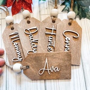 May include: Five wooden gift tags with names written on them. The names are "Mommy", "Daddy", "Hunter", "Conn", and "Ava". The tags are strung together with twine and white wooden beads.