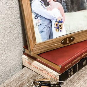Puede incluir: Una fotograf&iacute;a enmarcada de una pareja, apoyada en una pila de libros. Un llavero con el texto "FATHOR" y una definici&oacute;n est&aacute; en primer plano. Los libros son rojos y negros. El marco es de madera.