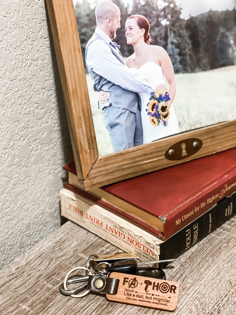 Puede incluir: Una fotograf&iacute;a enmarcada de una pareja, apoyada en una pila de libros. Un llavero con el texto "FATHOR" y una definici&oacute;n est&aacute; en primer plano. Los libros son rojos y negros. El marco es de madera.