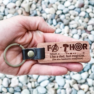 Puede incluir: Un llavero de madera con la palabra "FATHOR" grabada, junto con una definici&oacute;n. El llavero tiene una correa de cuero y un anillo de metal. El texto dice "Como un padre, pero m&aacute;s poderoso."