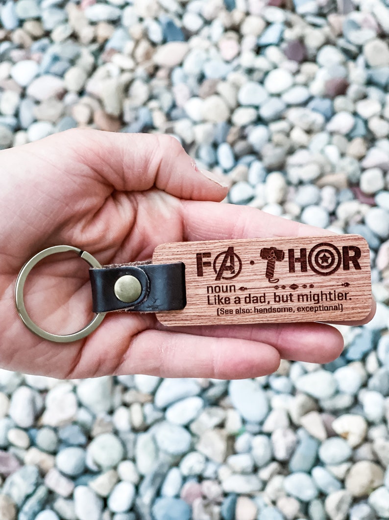 Puede incluir: Un llavero de madera con la palabra "FATHOR" grabada, junto con una definici&oacute;n. El llavero tiene una correa de cuero y un anillo de metal. El texto dice "Como un padre, pero m&aacute;s poderoso."
