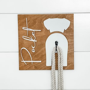 May include: A wooden dog leash holder with the word "Pocket" written in white lettering. The holder is shaped like a dog with a hook for hanging a leash. A white rope leash is hanging from the hook.
