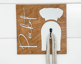 May include: A wooden dog leash holder with the word "Pocket" written in white lettering. The holder is shaped like a dog with a hook for hanging a leash. A white rope leash is hanging from the hook.
