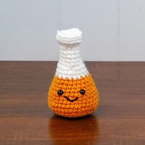 May include: A small, handmade crochet amigurumi in the shape of a laboratory flask. The base is bright orange with a stitched black smiling face and two small black bead eyes, while the narrow neck is crafted from white yarn.