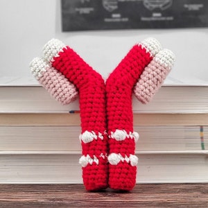 Puede incluir: Un juguete de amigurumi de color rojo y blanco en forma de Y con detalles blancos. El juguete está sentado sobre una pila de libros.