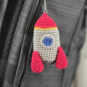 May include: A crocheted rocket-shaped keychain in shades of red, gray, yellow, and blue. The rocket has a red top, gray body, yellow trim, and red fins. A blue circle represents the window. The keychain is attached to a silver chain.