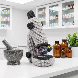 May include: A handcrafted crochet microscope featuring a light gray body with dark gray accents on the knobs, base, and eyepiece. The textile sculpture mimics the structure of a laboratory microscope with a vertical stand, stage, and objective lenses.