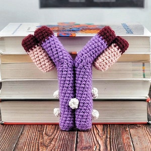 Puede incluir: Un juguete de amigurumi de color morado y rosa tejido a crochet con forma de cromosoma Y. El juguete está sentado encima de una pila de libros.