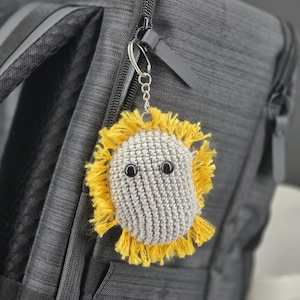 Puede incluir: Un llavero de ganchillo hecho a mano con una cara gris, ojos de botón negros y una franja amarilla. El llavero está sujeto a una cadena plateada y a un tirador de cremallera negro en una mochila gris.