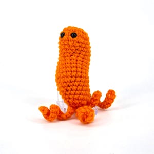 May include: A bright orange crochet creature with black eyes and curly tentacles. This handmade amigurumi is a whimsical, soft sculpture, suitable as a child's toy or a unique decorative item. The plushie is made with a tight stitch.