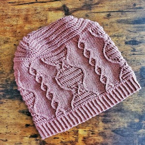 May include: A dusty rose knitted beanie featuring a prominent central cable knit pattern flanked by vertical twisted rope textures. The hat is constructed with a ribbed brim at the base and a soft, textured yarn finish throughout the entire garment.