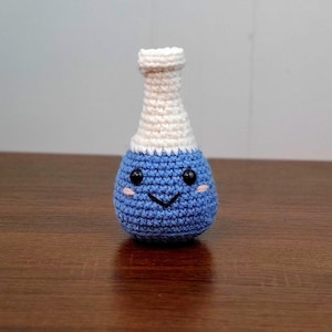 May include: A small, handmade crochet amigurumi toy shaped like a potion bottle. The base is round and blue with a stitched black V-shaped mouth, two black bead eyes, and small pink blush marks. The neck of the bottle is narrow and white.