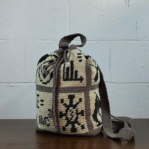 May include: A cylindrical crochet drawstring bag featuring a cream-colored base with dark brown geometric patterns and symbols. The bag includes a solid brown crocheted top border, a matching drawstring closure, and a long, flat crocheted shoulder strap.