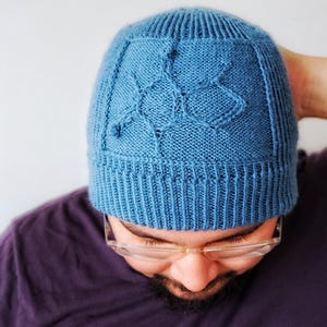 May include: A blue knitted beanie featuring a ribbed brim and a textured geometric pattern on the crown. The hat is constructed with a stockinette stitch body and a vertical ribbed cuff, showcasing a subtle, raised design integrated into the knit.