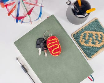May include: A red crocheted keychain shaped like a hot dog with a yellow mustard drizzle pattern, attached to a silver metal keyring with two black keys. The keychain rests on a sage green notebook next to a pen, a pencil holder, and a coaster.