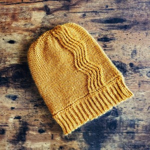 May include: A mustard yellow knit beanie featuring a ribbed brim and a vertical wavy cable knit pattern extending from the crown to the edge. The hat is constructed with a thick yarn texture and displays a consistent golden hue throughout the design.