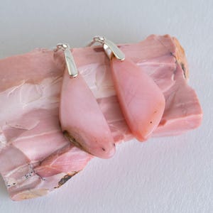 May include: A pair of teardrop-shaped earrings with a pale pink hue. The earrings have a silver-colored top and are displayed on a pink, textured surface. The earrings are a jewelry item.