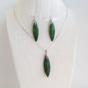Könnte beinhalten: Ein silbernes Halsketten- und Ohrring-Set mit grünen, blattartigen Anhängern. Die Halskette hat eine zarte Kette und die Anhänger haben ein silbernes Detail oben.