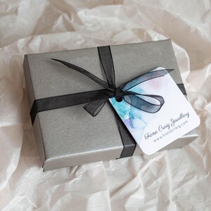K&ouml;nnte beinhalten: Eine graue Geschenkbox mit einem schwarzen Band und einem wei&szlig;en Etikett mit der Aufschrift "Fiona Craig Jewellery www.fionacraig.com"