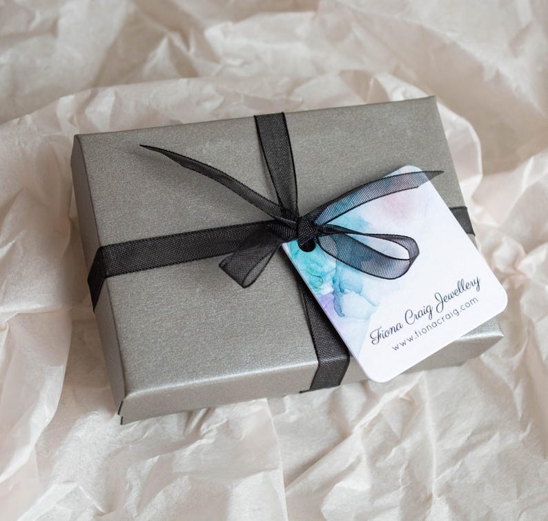 K&ouml;nnte beinhalten: Eine graue Geschenkbox mit einem schwarzen Band und einem wei&szlig;en Etikett mit der Aufschrift "Fiona Craig Jewellery www.fionacraig.com"