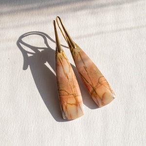 May include: Dangle earrings with a teardrop shape. The earrings have a light peach and cream stone with brown veining. The ear wires are gold-coloured metal. The earrings are displayed on a white surface.