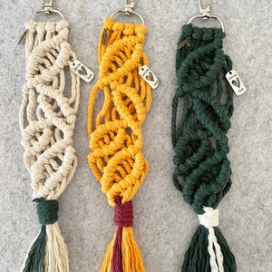 May include: Three macrame keychains with a silver metal clasp and a tassel. The keychains are in cream, yellow, and green. Each keychain has a silver charm with the text "Follow Your" and a heart.