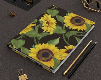 May include: A dark brown notebook with a sunflower pattern. The sunflowers are yellow with brown centers and green leaves. The notebook is closed and lying on a dark brown surface.