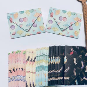 Puede incluir: Dos sobres pequeños y cuadrados con un fondo azul claro y un patrón de macarons, abanicos y otros elementos decorativos. Debajo hay varias pilas de tarjetas plegadas con varios patrones, incluyendo zapatos y otros diseños.