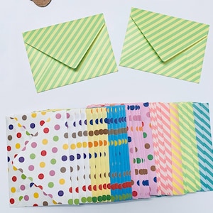 Puede incluir: Una colección de sobres estampados de colores. En la parte superior, dos sobres con rayas diagonales verdes y blancas. Debajo, sobres con lunares y rayas en varios colores, incluyendo rosa, amarillo y azul.