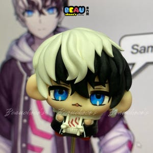 May include: A small clay figurine of a character with split-coloured hair, featuring white on the left and black on the right. The figure has large, bright blue eyes, a small mouth, and wears a black and white hooded jacket with red accents.