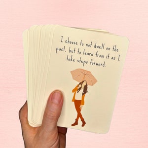May include: A stack of cards with a quote about learning from the past and moving forward. The quote reads "I choose to not dwell on the past, but to learn from it as I take steps forward." An illustration of a person holding an umbrella is on the card.