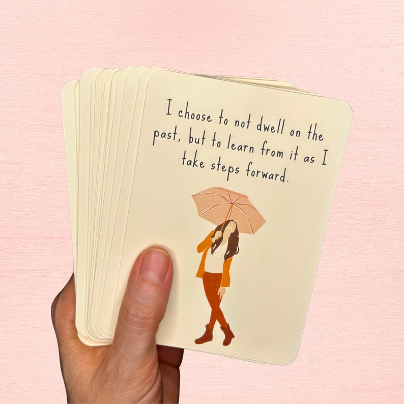 May include: A stack of cards with a quote about learning from the past and moving forward. The quote reads "I choose to not dwell on the past, but to learn from it as I take steps forward." An illustration of a person holding an umbrella is on the card.