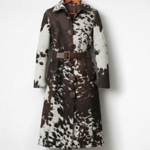 May include: A long, cowhide coat with a brown and white spotted pattern. The coat features a notched collar, long sleeves, a brown belt, and button closures. The coat is hanging on a wooden hanger.