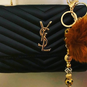 Puede incluir: Un bolso negro acolchado con un logotipo YSL dorado y una correa de cadena dorada. Un llavero dorado con un pompón de piel marrón y cuentas doradas cuelga del bolso.