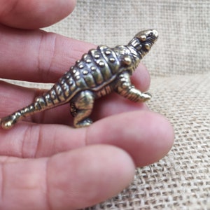 Może przedstawiać: Mała, szczegółowa figurka dinozaura z mosiądzu, prawdopodobnie Ankylozaura, z teksturowaną skórą i długim ogonem. Figurka jest trzymana w dłoni, ukazując jej rozmiar i skomplikowany design. Dinozaur ma złoto-brązowy kolor.