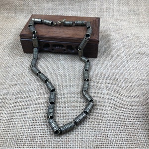 Könnte beinhalten: Eine silberfarbene Halskette mit zylindrischen Perlen mit einem sich wiederholenden geometrischen Muster. Die Halskette wird auf einer neutralen Oberfläche mit einer Holzkiste im Hintergrund präsentiert.