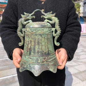 May include: An antique bronze bell with a patina finish. Two dragon figures form the handle. The bell has intricate carvings and Chinese characters. The base has a flared design. The bell is held by a person.