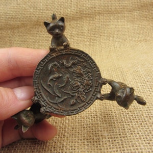 Puede incluir: Un objeto decorativo de color bronce con tres patas y tres figuras de gato. Cada gato está posicionado alrededor de una pieza central circular con un diseño floral y de dragón en relieve. El objeto está sostenido por una mano.