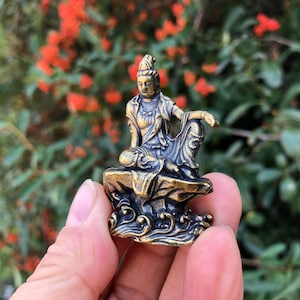 Può includere: Piccola statua color bronzo di una figura seduta con dettagli ornati. La figura è seduta su una base strutturata con motivi a vortice. La statua ha un'espressione serena ed è ambientata su uno sfondo sfocato di fogliame verde e fiori rossi.